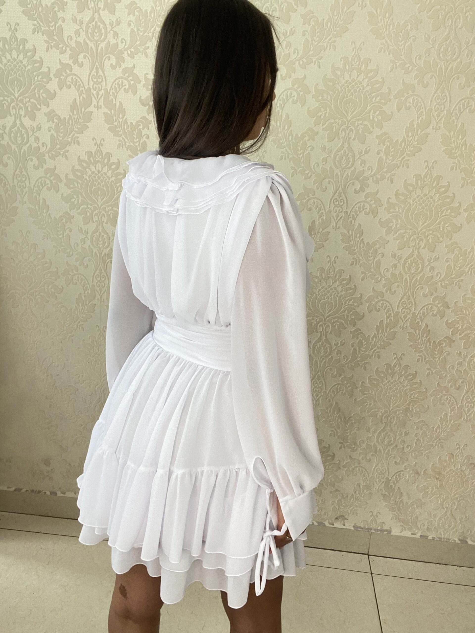 Back view of Sophia White Mini Party Dress showcasing a fitted waist, flowing layers, and delicate long sleeves.